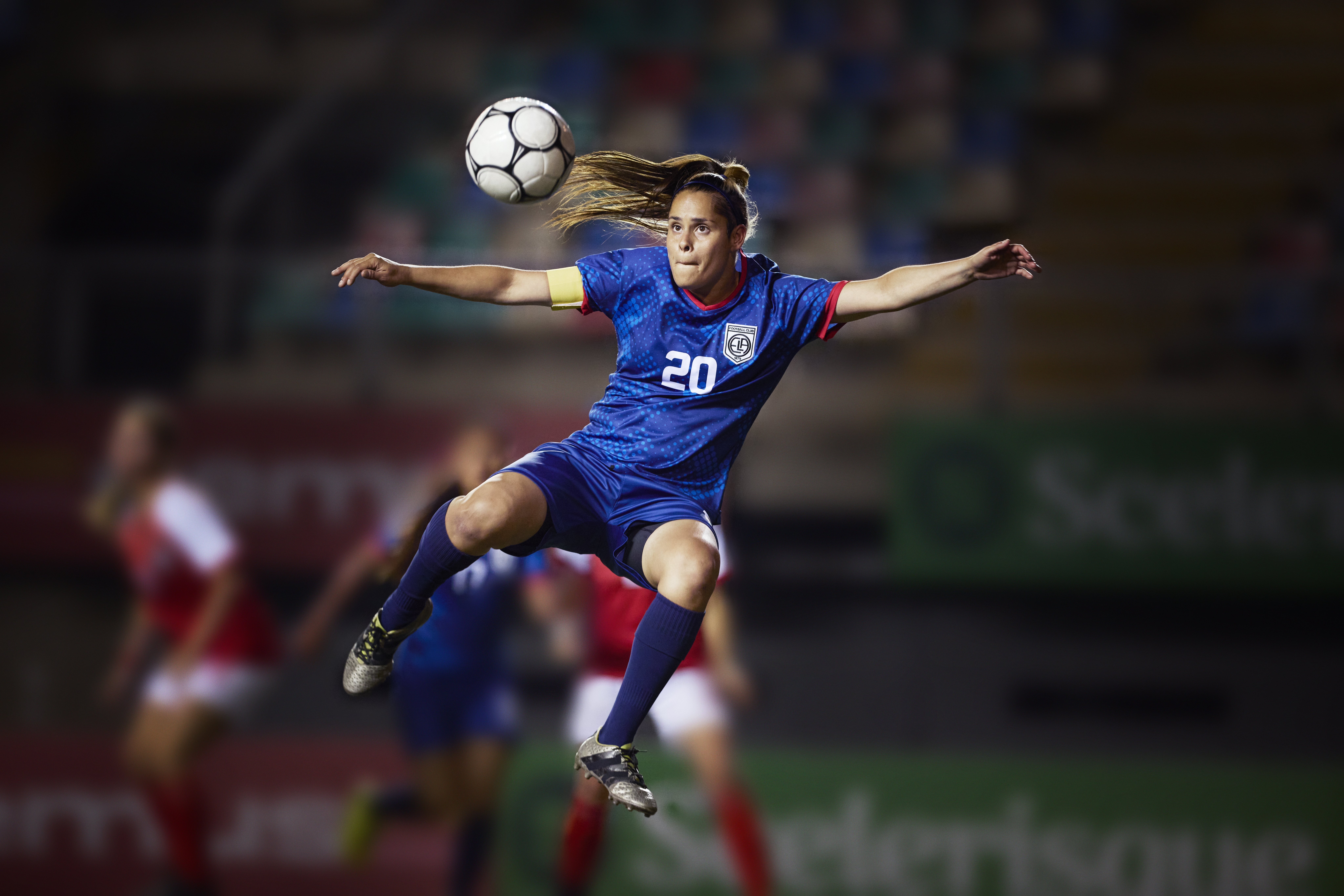 Action shot of a serious professional female soccer player that is focusing on the soccer ball in preparation to strike it in mid-air into the goal. Female soccer player with brown hair and a royal blue uniform with the number 20 on the jersey.
