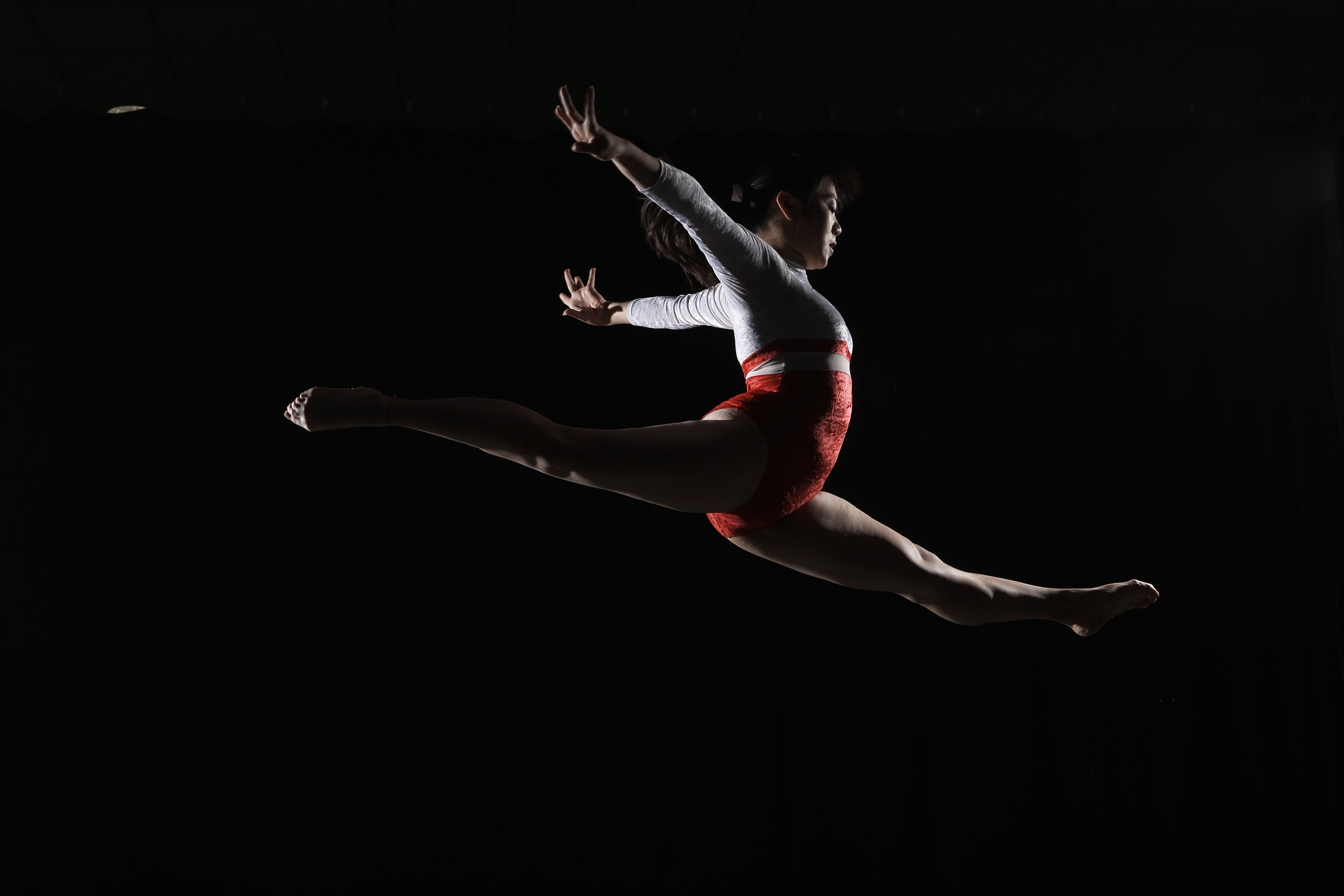 Young female gymnast leaping in mid-air, arms outstretched, side view. Female with dark brown hair and a red and white gymnastics uniform.