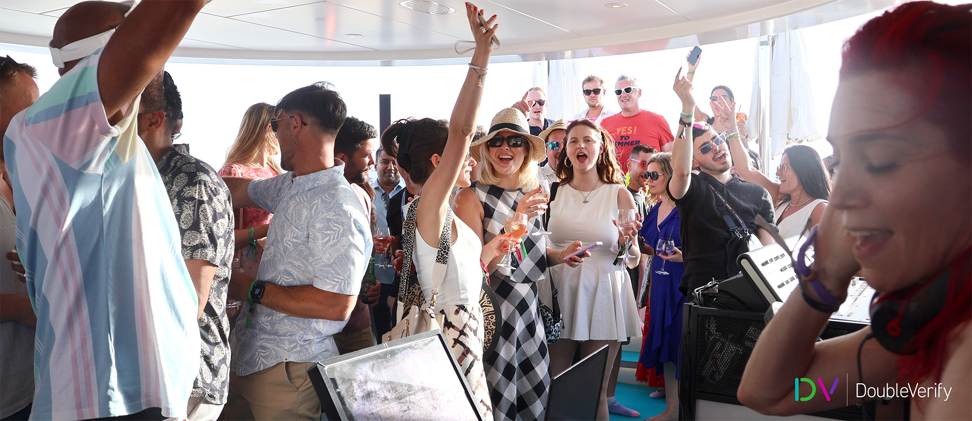 Photo taken on DV's yacht at the 2024 Cannes Lions International Festival of Creativity showing a large group of guests dancing and laughing with drinks in their hands. In the corner, you can see the DJ holding headphones to her ear.